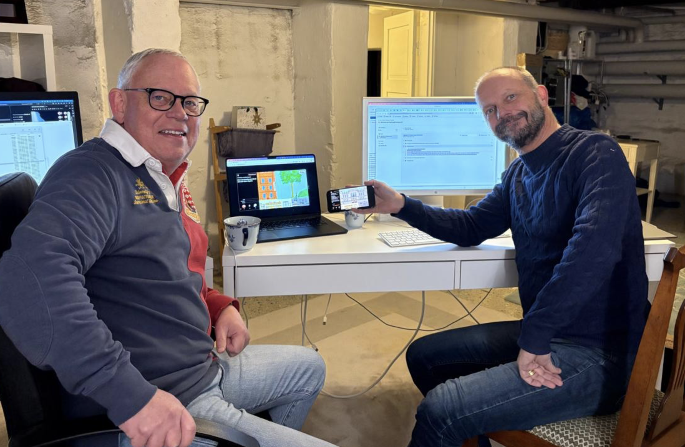 Stefan Wiklund och Anders Bjerkén sitter vid ett skrivbord i kodgrottan 2026 och arbetar med remaken av Dubbelmoral! – med laptop, telefon och stor skärm.